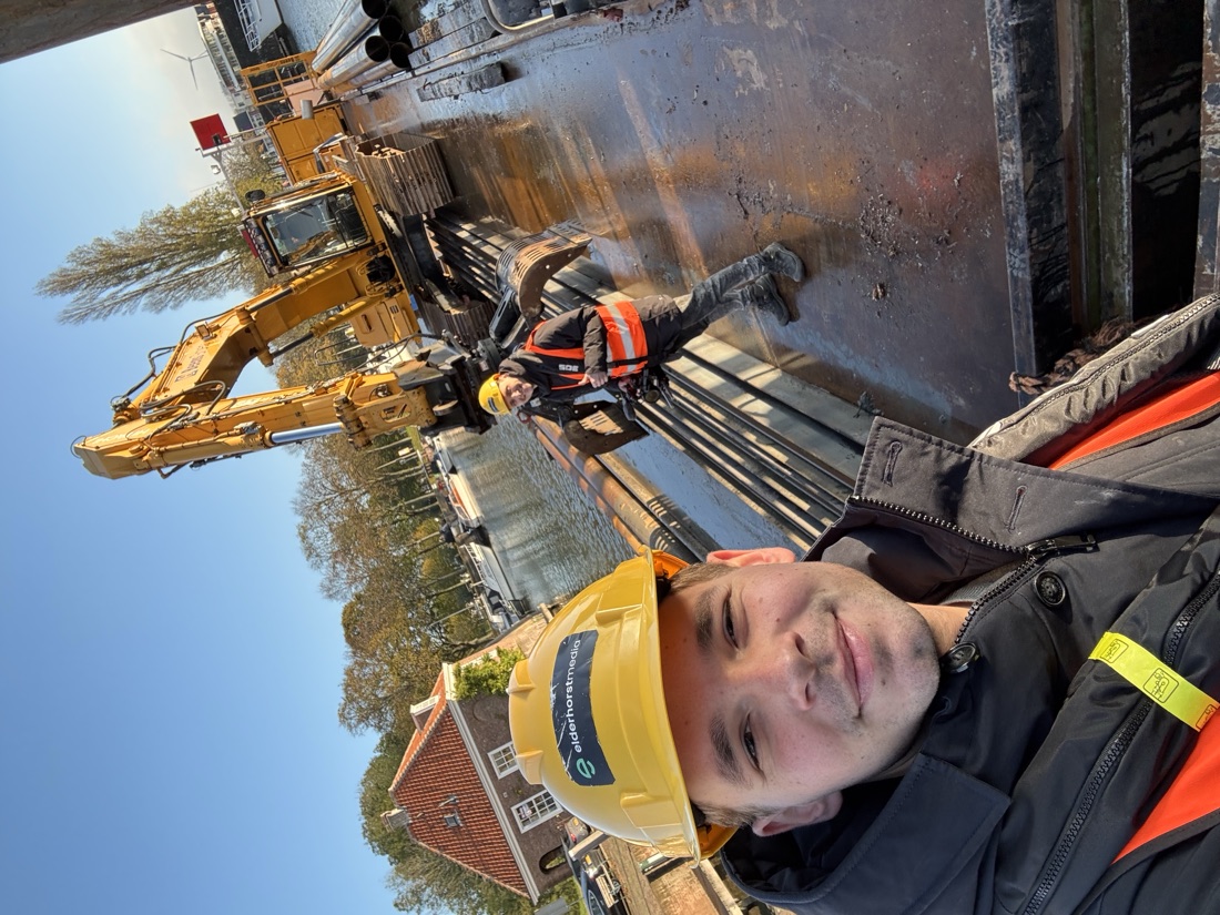 Stan Elderhorst met een Elderhorst Media-helm op een werklocatie bij het water, met een graafmachine en collega op de achtergrond.